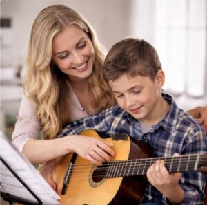 Klasik gitar çalmayı öğrenen bir çocuğa gülümseyerek eşlik eden kadın öğretmen